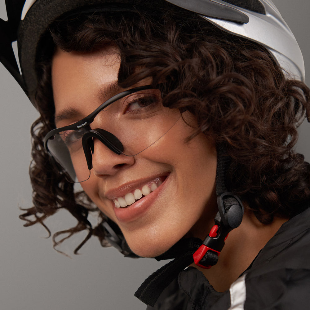 A person with curly hair smiles while wearing the Reebok Octane Shift bike helmet, smart eyewear featuring black and clear sports glasses, and a black jacket. The Reebok helmet strap is securely fastened under their chin.