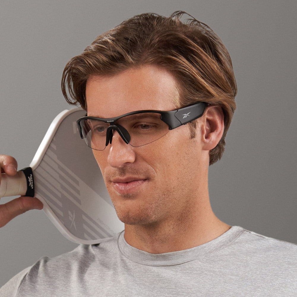 A man wearing Reebok Octane Shift smart eyewear with photochromic lenses and a light gray shirt holds a white pickleball paddle near his face, smiling slightly against a plain gray background.