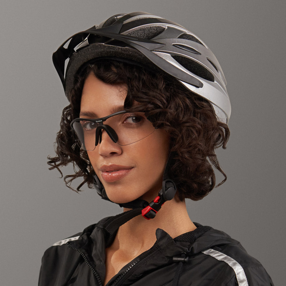 A woman with curly hair wears the Reebok Octane Shift smart eyewear with clear photochromic lenses, a silver bike helmet, and a black jacket, looking at the camera against a plain gray background.