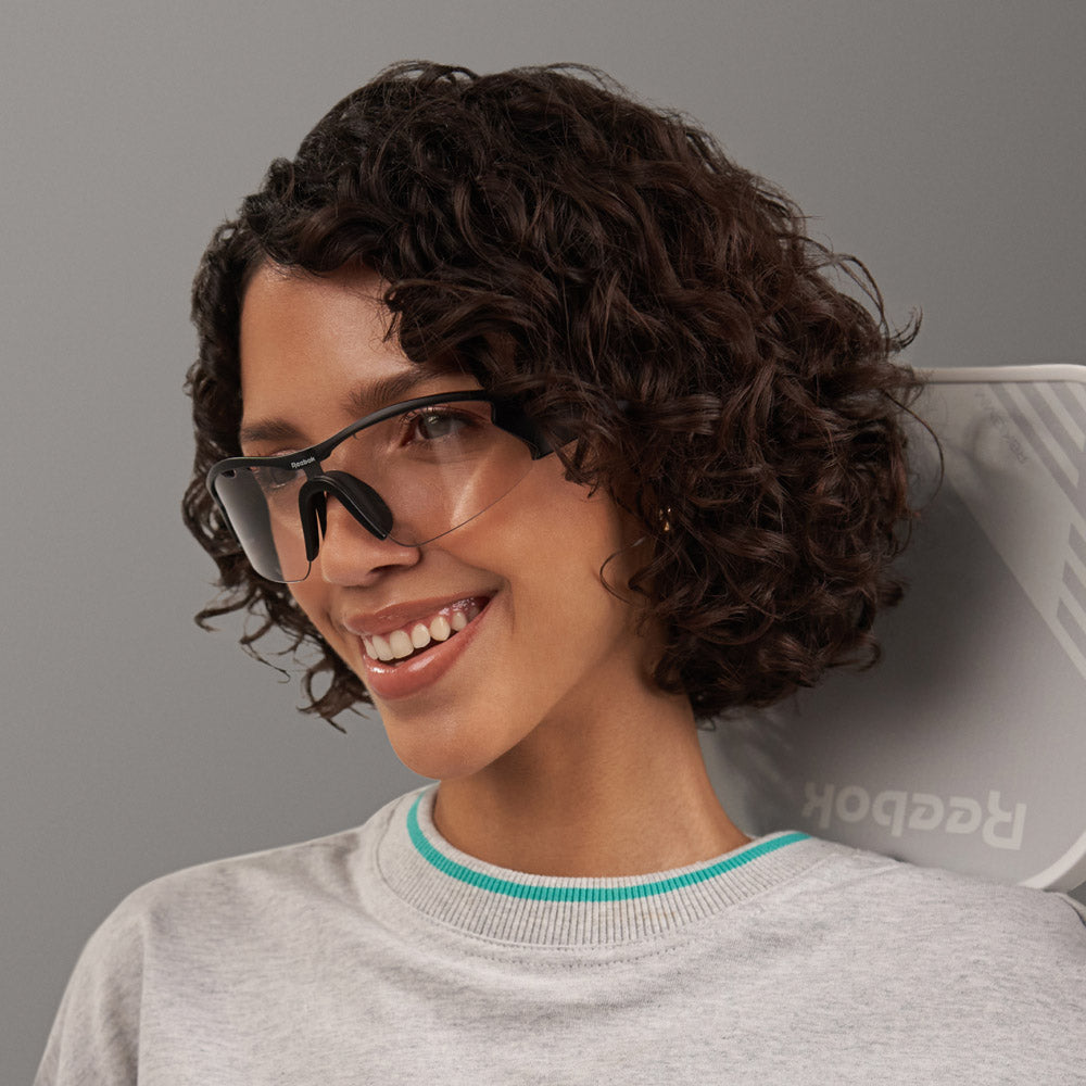 A smiling woman with short, curly dark hair wears large, transparent smart eyewear and a light gray sweatshirt. She stands against a plain gray background with a white Reebok Octane Shift behind her.