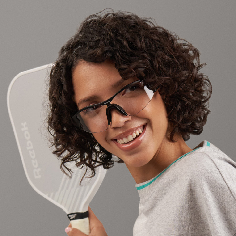 A young person with curly hair, wearing smart photochromic eyewear and a grey shirt, smiles while holding a Reebok Octane Shift paddle against a neutral grey background.