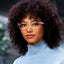 A confident woman with curly dark hair models Reebok's Shine smart glasses, paired with a light blue turtleneck. She looks directly at the camera against a softly blurred backdrop featuring hints of greenery.