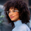 A woman with curly hair, wearing Reebok's Shine privacy-focused smart glasses and a light blue turtleneck sweater, smiles softly while gazing to the side. The background is gently blurred.