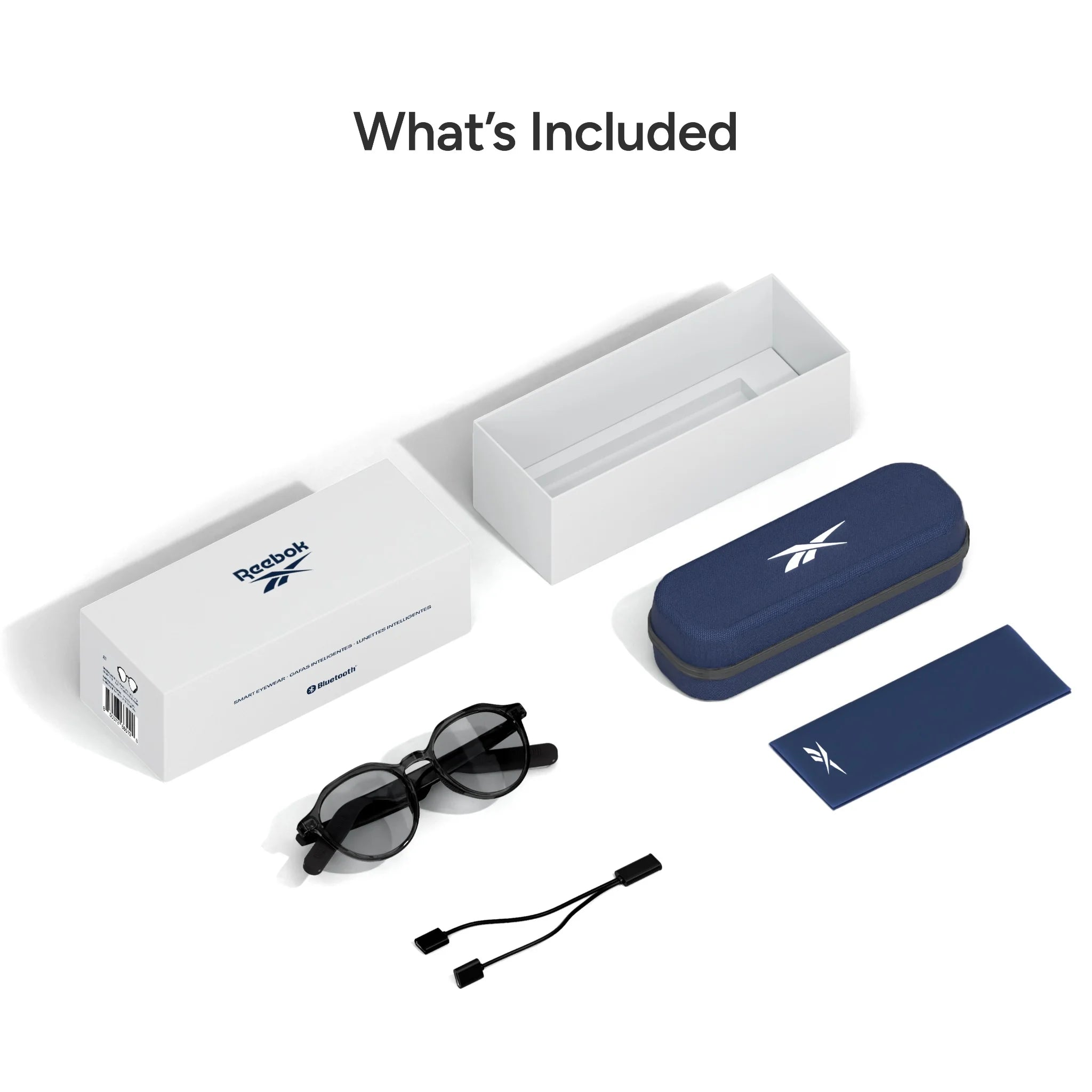Flat lay of Reebok Thunder smart glasses set showing a white box, navy blue hard case, open-ear audio sunglasses, blue cleaning cloth, and dual-ended USB cable with “What’s Included” above the items.