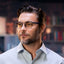 A man with short dark hair, a trimmed beard, and glasses is wearing the Reebok Thunder light blue collared shirt, looking slightly to the side in smart glasses with blurred bookshelves in the background.