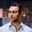 A man with short dark hair, a beard, and glasses, dressed in a light blue shirt, looks to the side while wearing Reebok Thunder smart glasses. Behind him are softly blurred bookshelves.