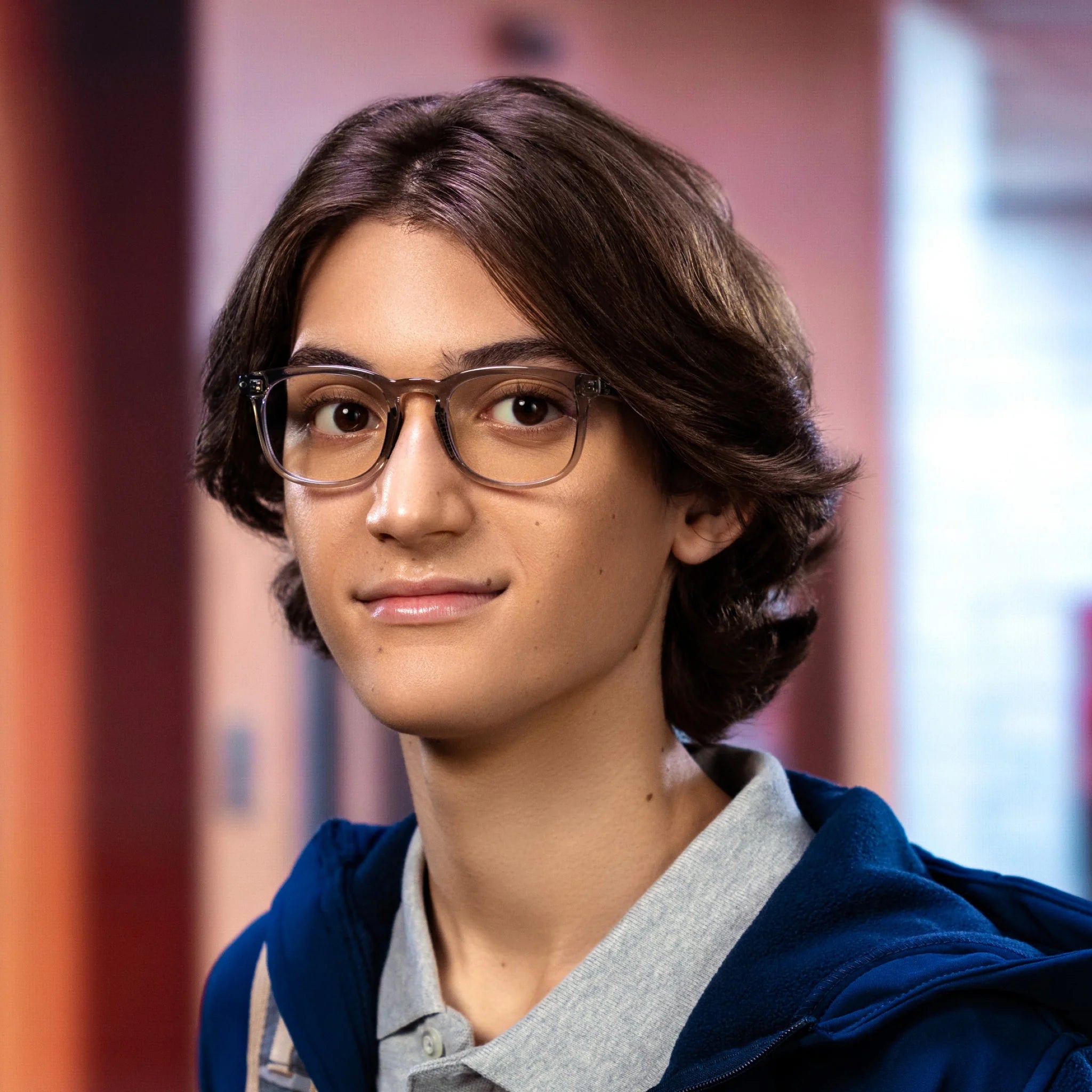 A young person with medium-length brown hair and glasses, wearing a blue hoodie over a light gray collared shirt, stands indoors against a soft backdrop, showcasing Reebok’s Thunder smart glasses featuring open-ear audio.