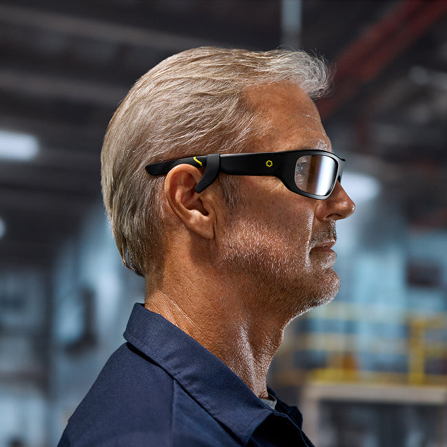 A middle-aged man with gray hair and stubble wears Lucyd Boosters bone conduction smart glasses with a black frame in an industrial setting, shown in right-facing profile. Designed for Lucyd Armor and Reebok.