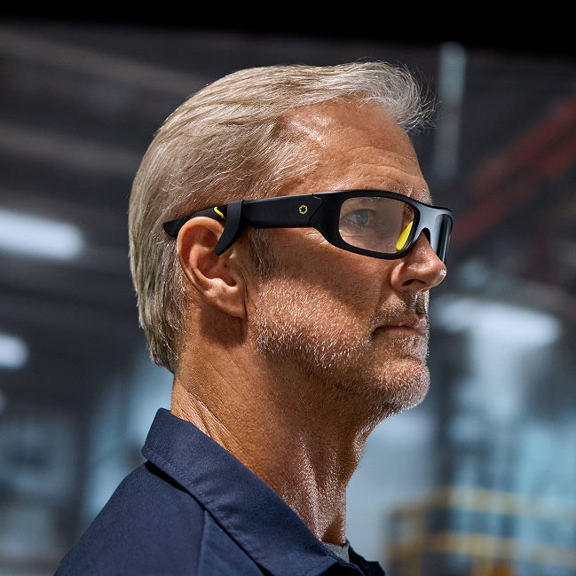 A mature man with short gray hair and a trimmed beard wears Lucyd Boosters smart glasses by Lucyd, featuring yellow accents, while standing indoors in an industrial or warehouse setting.