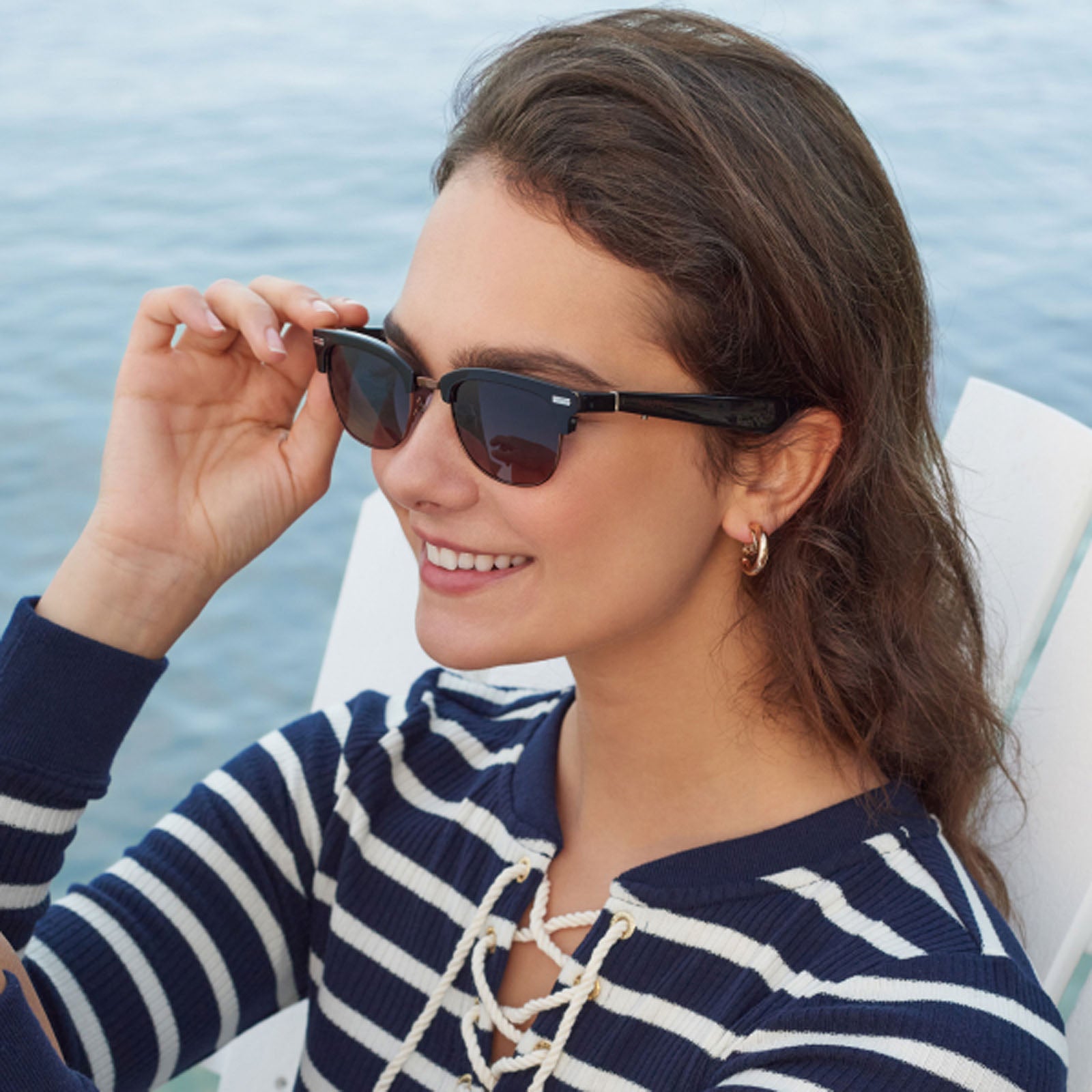 A woman wearing Nautica Spyglass sunglasses and a navy striped top sits on a white chair by the water, smiling and enjoying a sunny day.