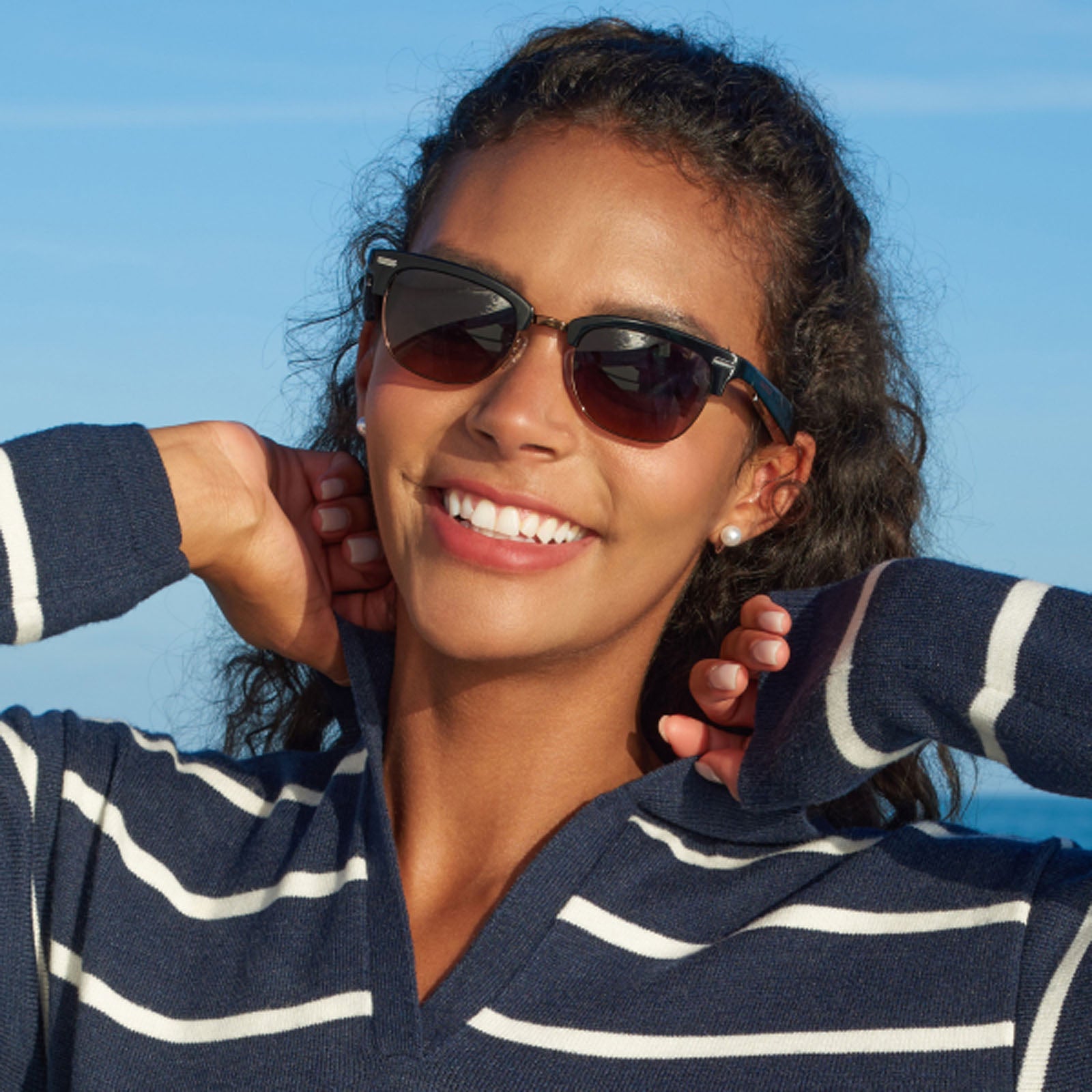 A smiling woman wears Nautica Spyglass Bluetooth 5.2 glasses and a navy-blue, white-striped sweater outdoors, pulling at her sweater’s neck with both hands under a clear blue sky.