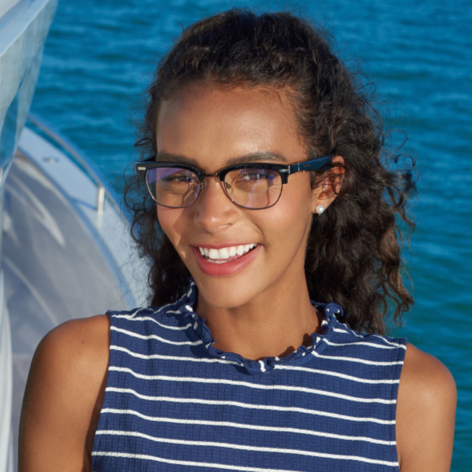 A smiling woman with curly hair and glasses stands on a boat in a blue and white striped sleeveless top, enjoying the sun in her Nautica Spyglass polarized UV400 clubmaster sunglasses, with sparkling blue water behind her.