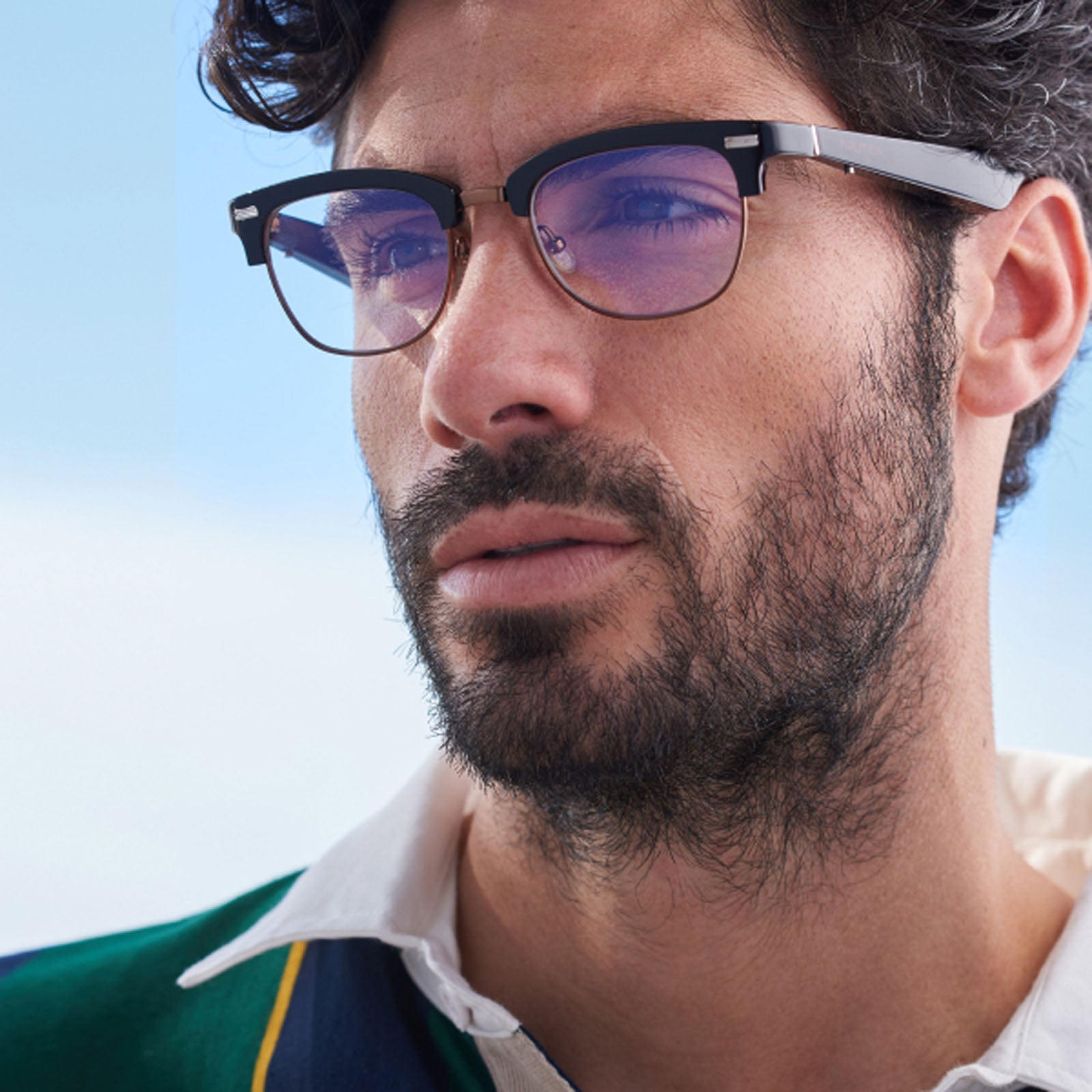 A man with dark hair, a beard, and glasses gazes thoughtfully into the distance. He wears a white-collared shirt, a colorful sweater, and Nautica Spyglass clubmaster sunglasses that stand out against the clear blue sky.
