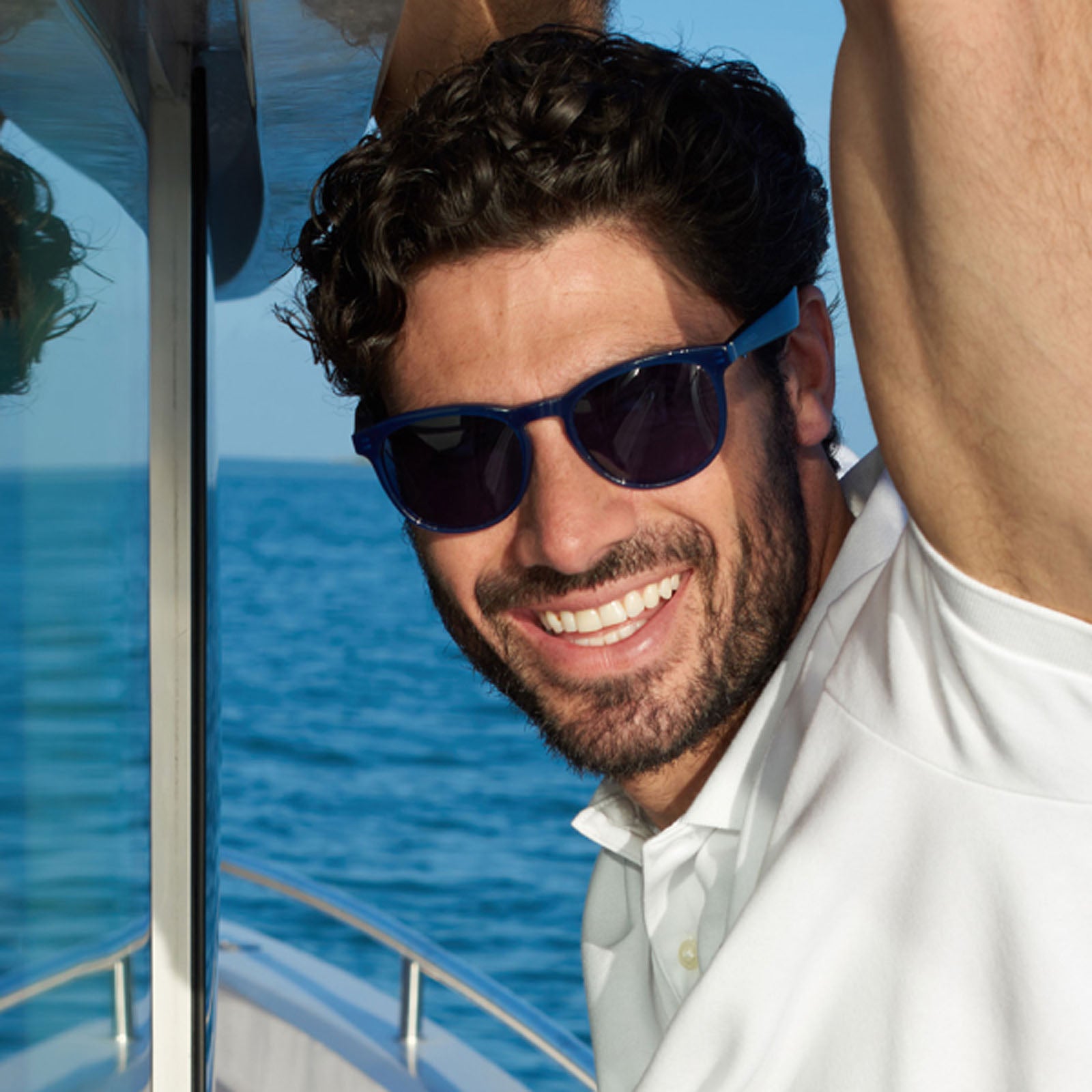 A man with curly hair and a beard, wearing Nautica Tailwind Bluetooth sunglasses and a white polo shirt, smiles while leaning against a boat railing with the ocean and clear blue sky in the background.