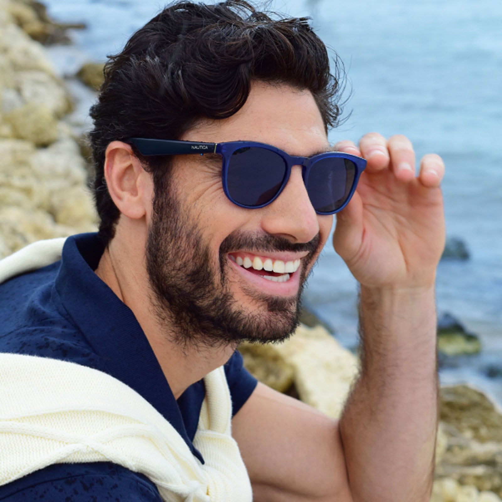 A smiling man with dark hair and a beard wears Nautica Tailwind polarized UV400 glasses and a navy shirt with a cream sweater over his shoulders, sitting on rocks by the sea.