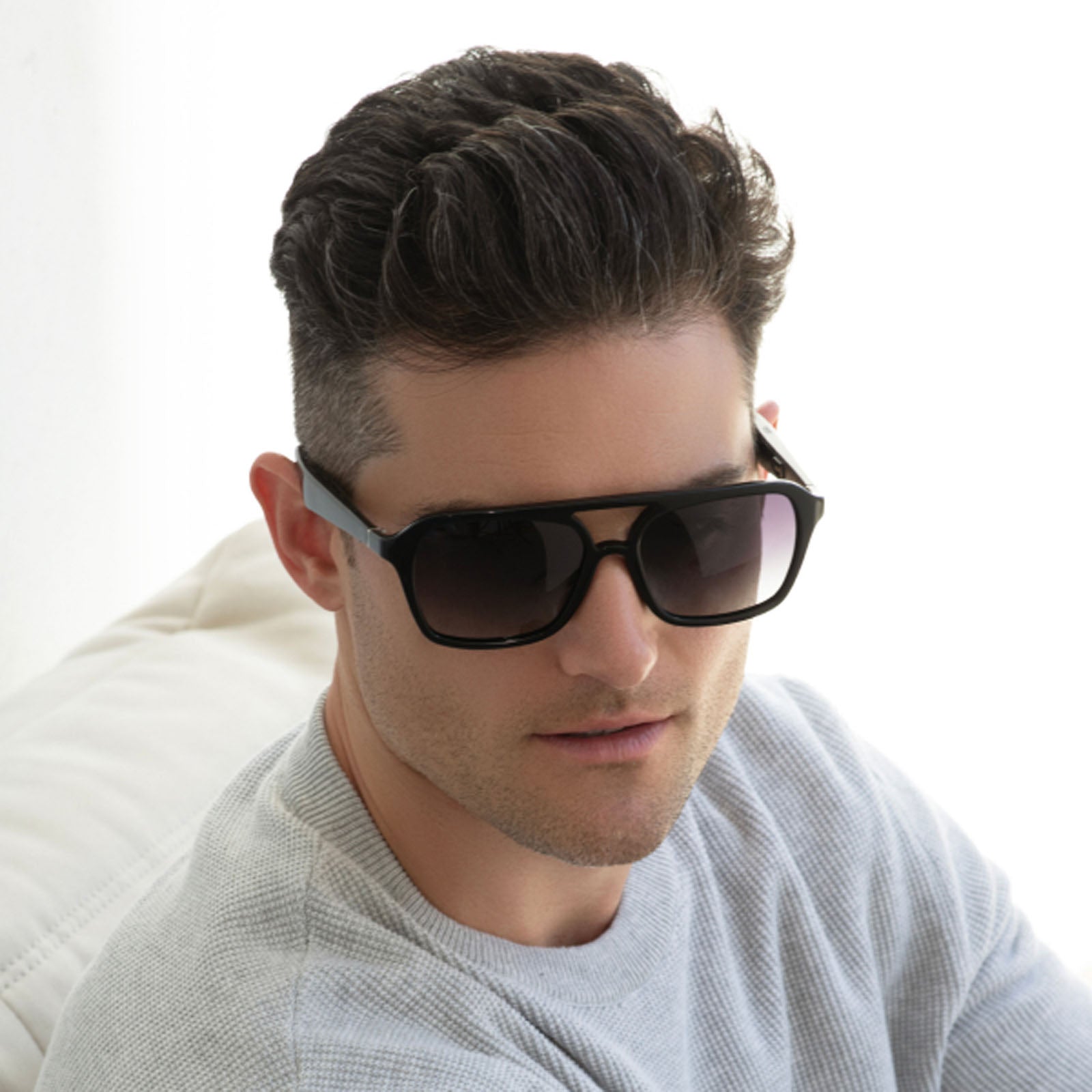 A man with short, dark hair is wearing Lucyd XL Voyager XXL large polarized UV protection glasses and a light gray sweater, sitting on a white textured surface against a bright background.