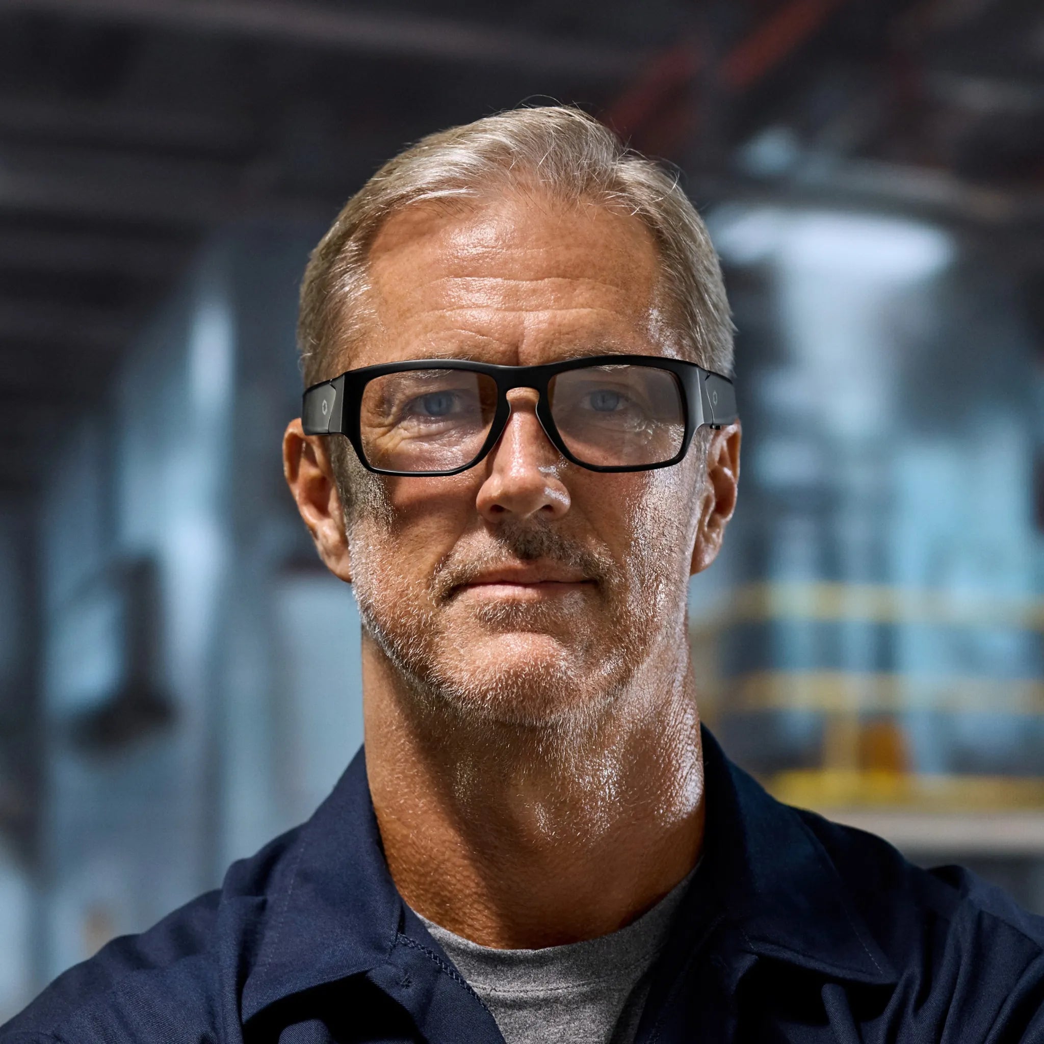 A middle-aged man with short gray hair and a beard confidently wears Lucyd Armor | Vantage Photochromic smart, prescription-compatible safety glasses by Lucyd in an industrial setting, with blurred machinery visible in the background.