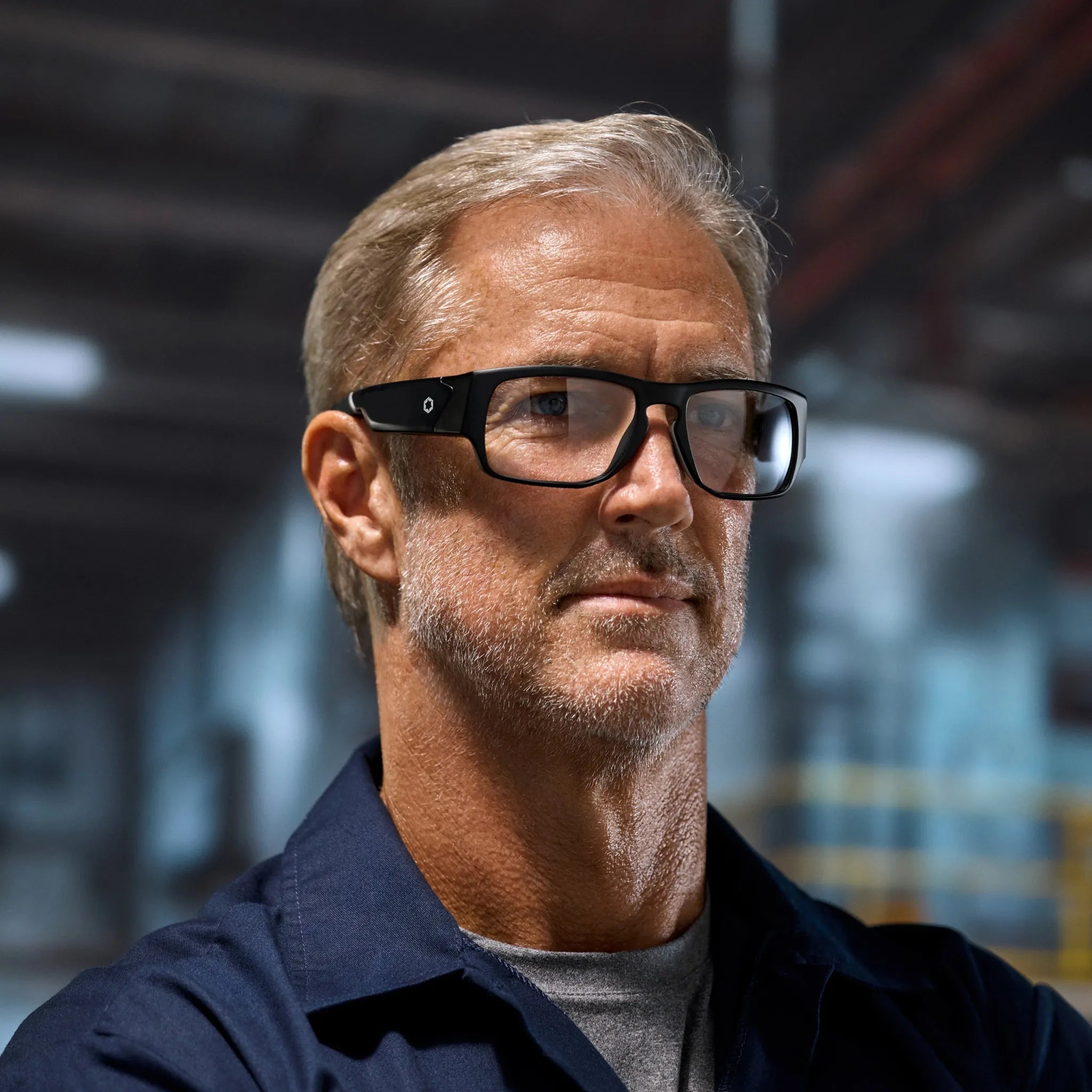 A middle-aged man with short gray hair and a trimmed beard wears Lucyd Armor | Vantage Photochromic smart safety glasses by Lucyd in a warehouse, dressed in a navy blue work shirt and gray t-shirt, looking confidently into the distance.