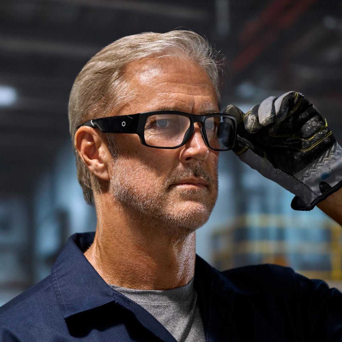 A middle-aged man with light hair, in a blue work shirt and glove, stands in an industrial warehouse, looking focused as he adjusts his Lucyd Armor | Vantage Photochromic smart safety glasses by Lucyd.