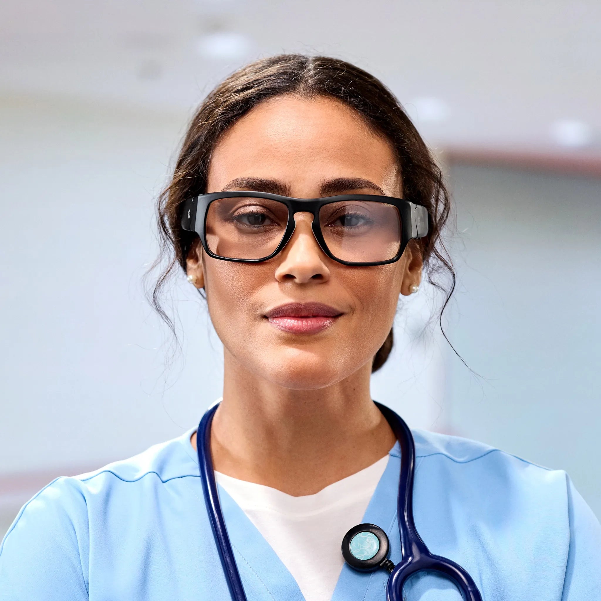 A woman in light blue scrubs wears Lucyd Armor | Vantage Photochromic smart safety glasses by Lucyd and a stethoscope, her dark hair pulled back and expression calm, standing indoors with a blurred background.