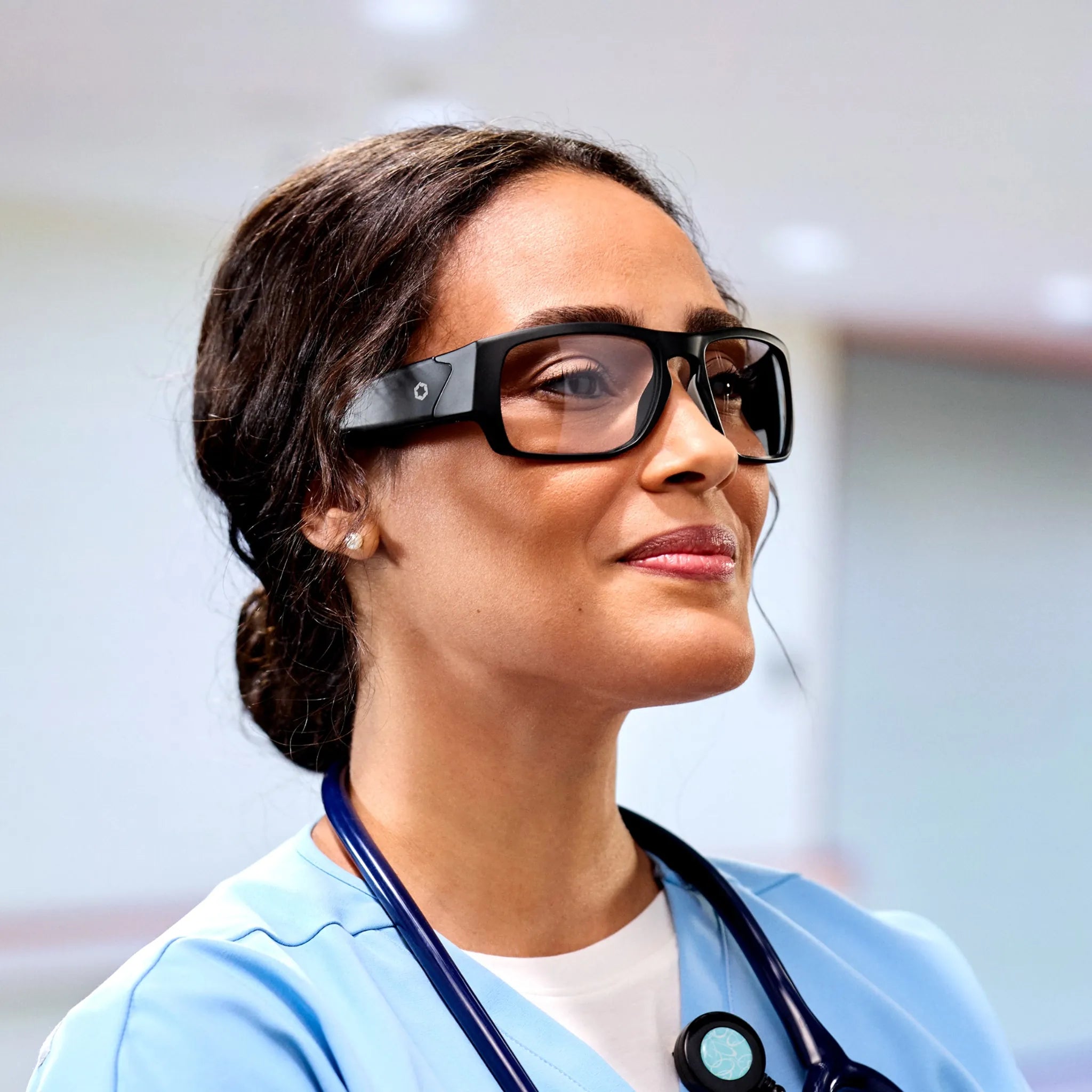 A woman in medical scrubs wears Lucyd Armor | Vantage Photochromic smart safety glasses by Lucyd. With her hair tied back and a stethoscope around her neck, she exudes confidence as a healthcare professional indoors.