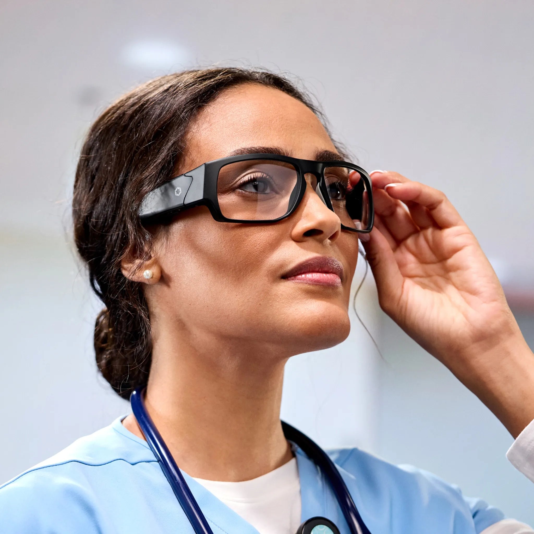 A healthcare professional in scrubs wears Lucyd Armor | Vantage Photochromic smart safety glasses with a focused expression, holding the frame, and a prescription-compatible stethoscope around her neck.
