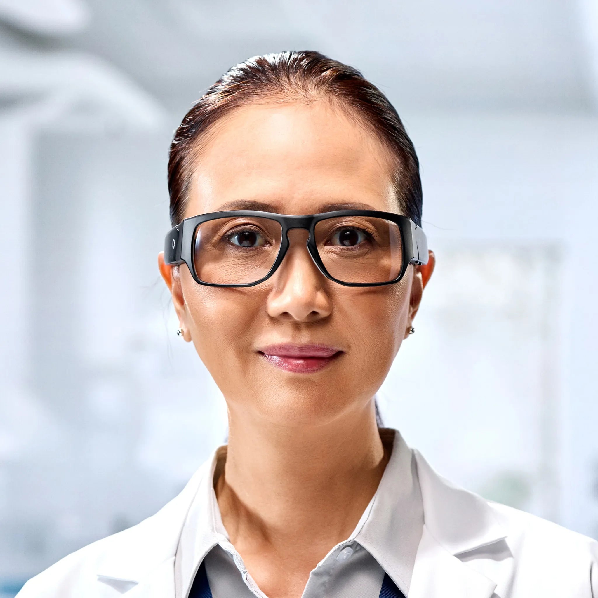 A woman wearing Lucyd Armor | Vantage Photochromic prescription-compatible safety glasses and a white lab coat stands confidently in a bright, modern laboratory.