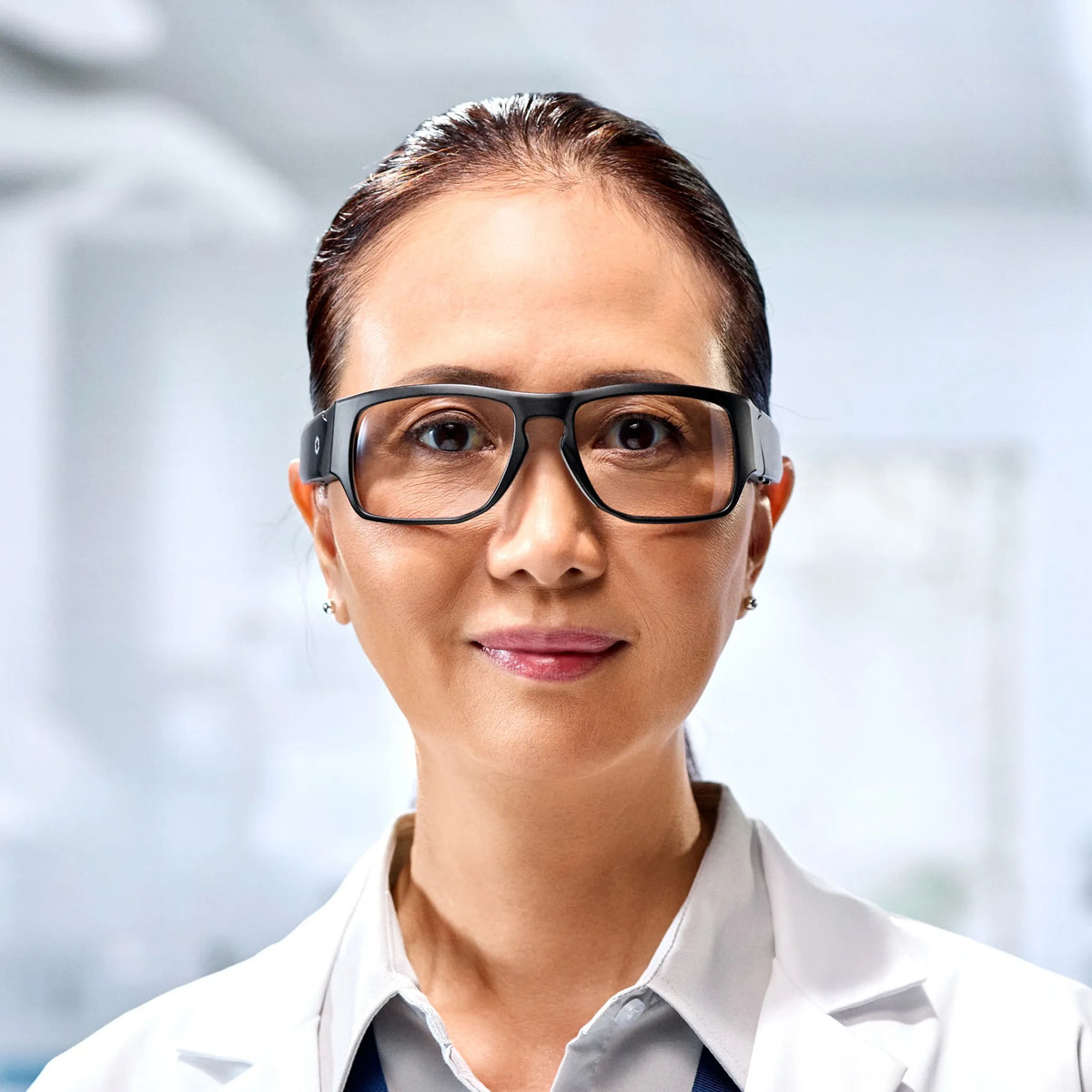 A woman wearing Lucyd Armor | Vantage Photochromic prescription-compatible safety glasses and a white lab coat stands confidently in a bright, modern laboratory.