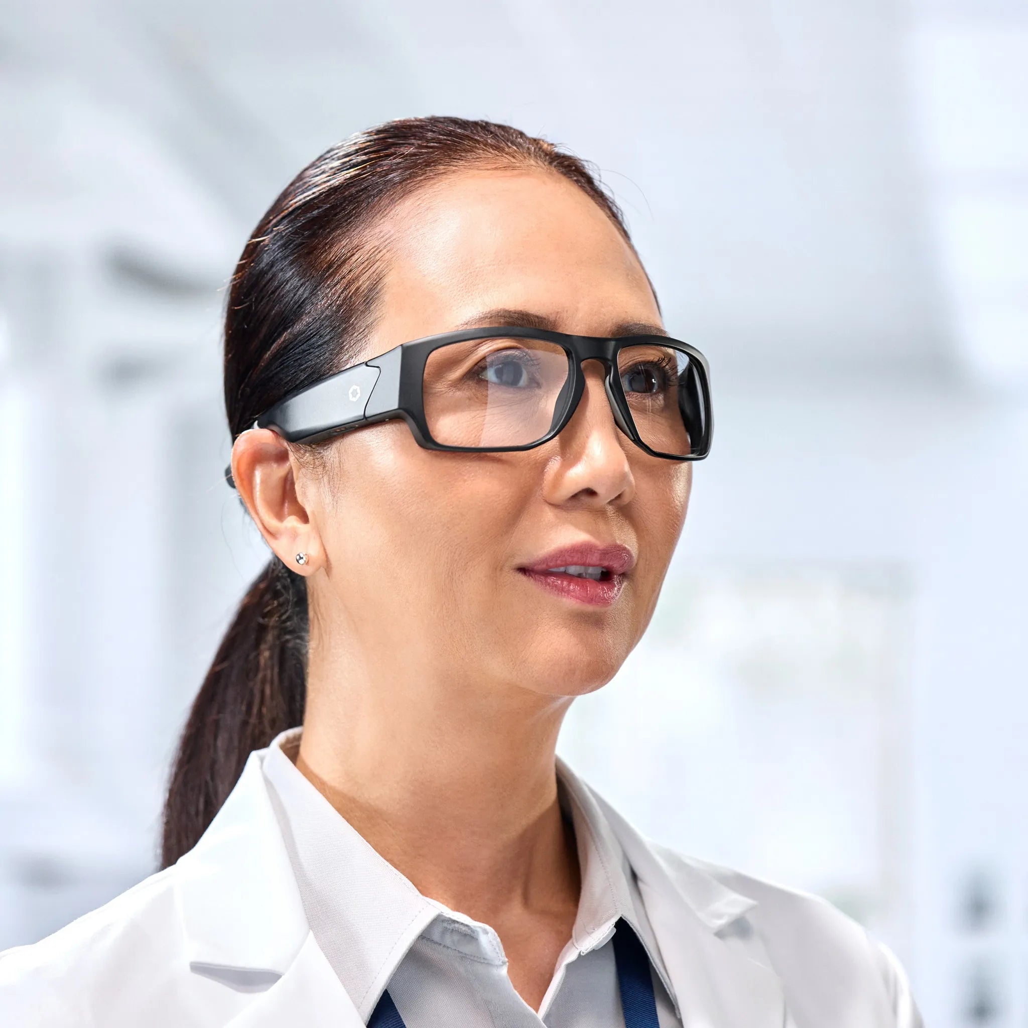A woman in a white lab coat wears Lucyd Armor | Vantage Photochromic smart safety glasses by Lucyd, looking ahead in a brightly lit, modern indoor lab or medical facility.